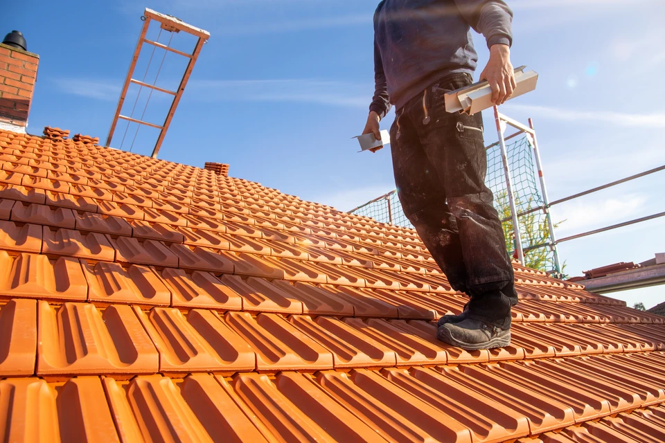 Photographie en ext&eacute;rieur d'un chantier de r&eacute;novation de toiture. Un ouvrier en tenue professionnelle travaille sur une toiture en cours de r&eacute;paration ou de remplacement, visible sur la structure d'un b&acirc;timent. La sc&egrave;ne montre clairement des travaux de couverture toiture (tuiles, ardoise ou mat&eacute;riau de couverture), avec un environnement de chantier organis&eacute; et professionnel en pleine lumi&egrave;re naturelle.